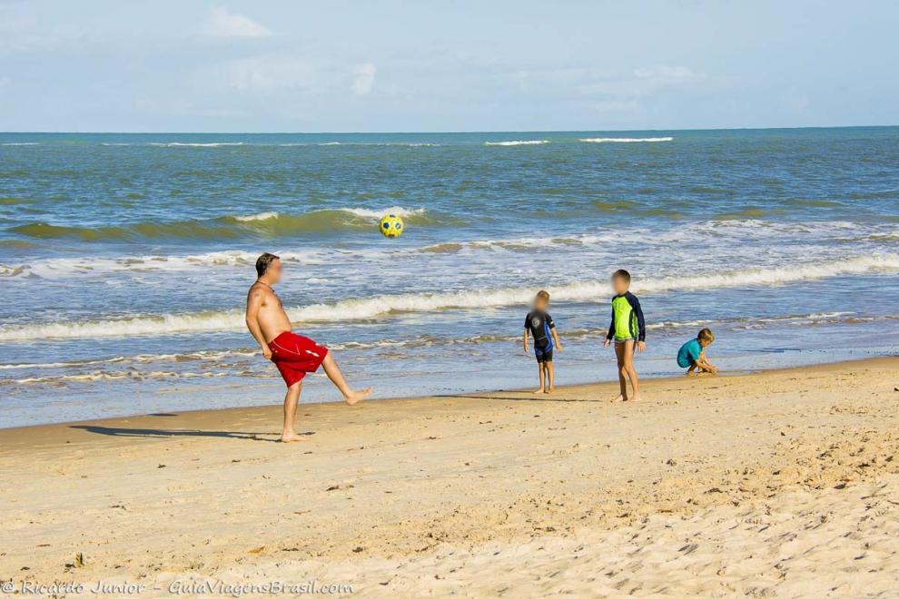 Imagem de um pai jogando bola com filhos na beira da praia.